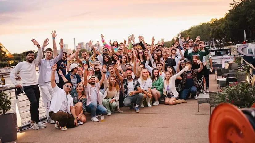 A large group of colleagues waving on an outdoor waterfront terrace at sunset, perfect for a team offsite.