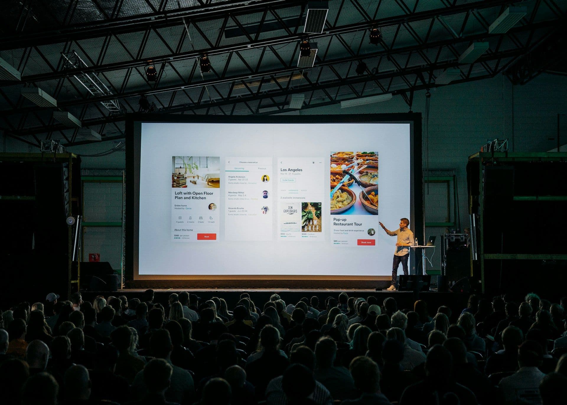 Man presents on a large screen to an audience at a corporate seminar in an industrial event space.