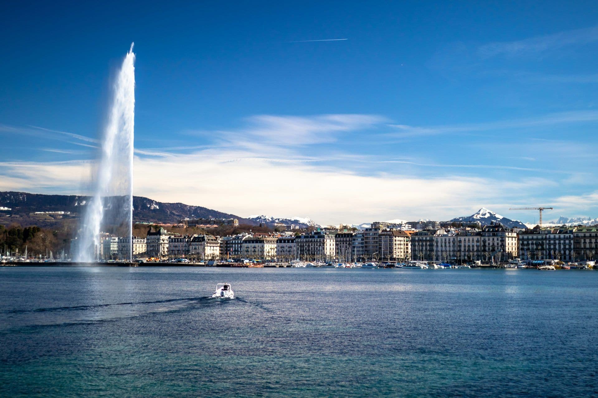 The iconic Jet d'Eau fountain sprays high over Lake Geneva with the city skyline and snowy mountains in the