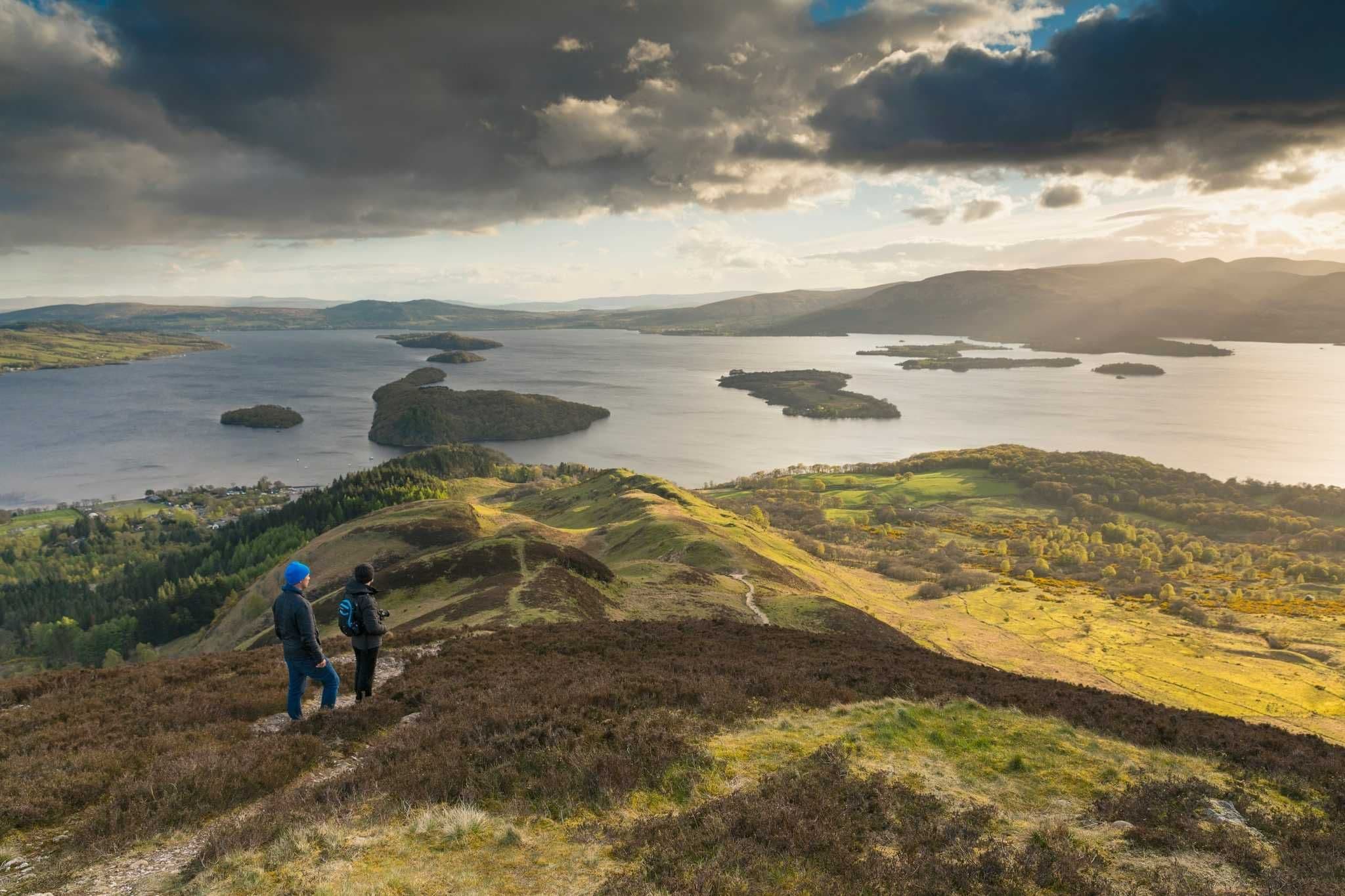 Two hikers on Conic Hill overlook Loch Lomond and its islands under a dramatic sky in Scotland.