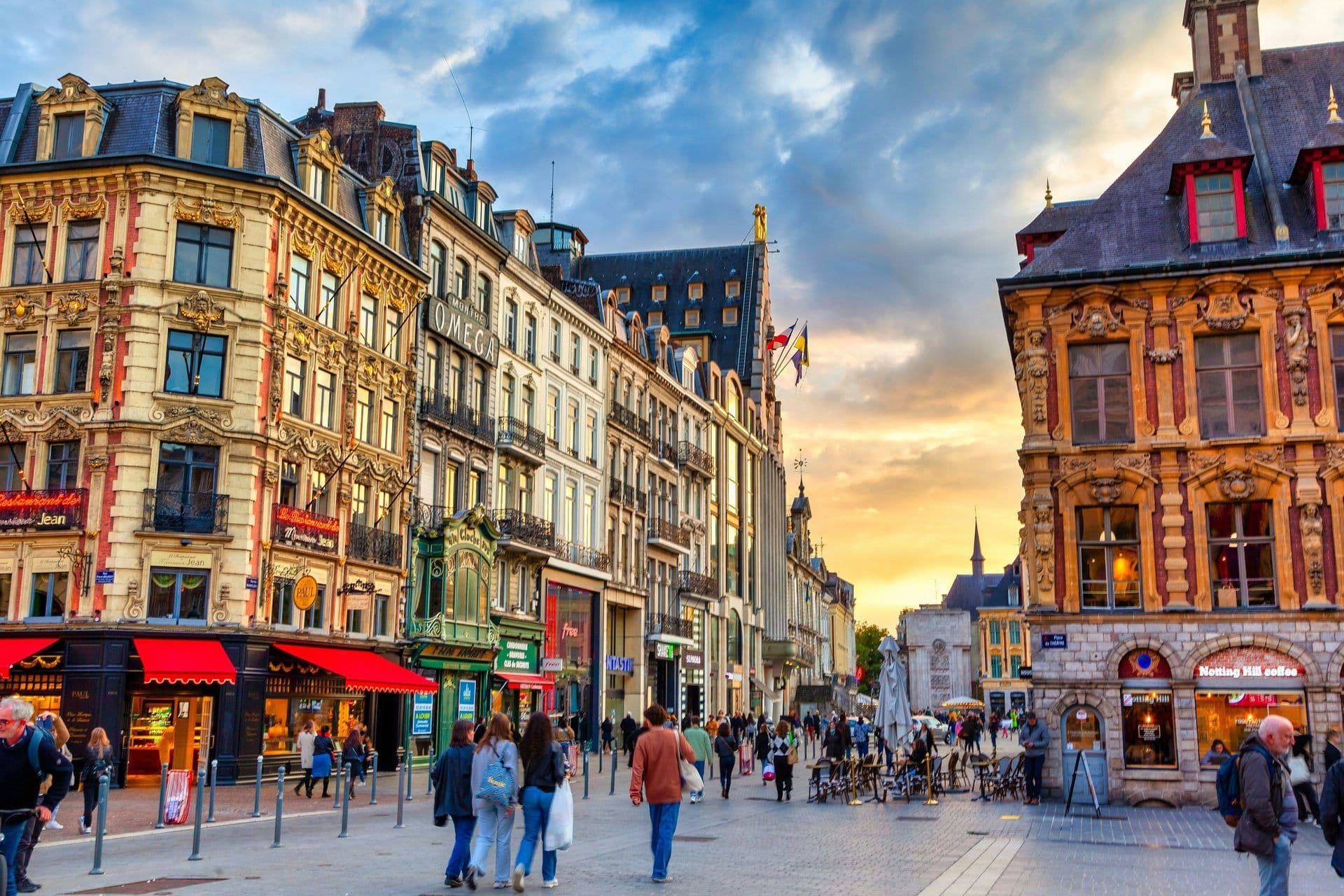 Historic buildings line a lively street in Lille, France, with shops, restaurants, and pedestrians at sunset.