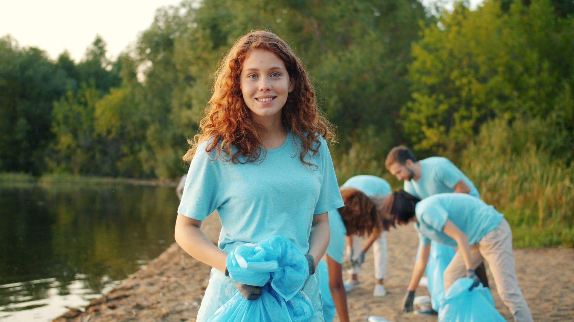 A smiling woman holds a trash bag during an outdoor team building clean-up event by a lake.