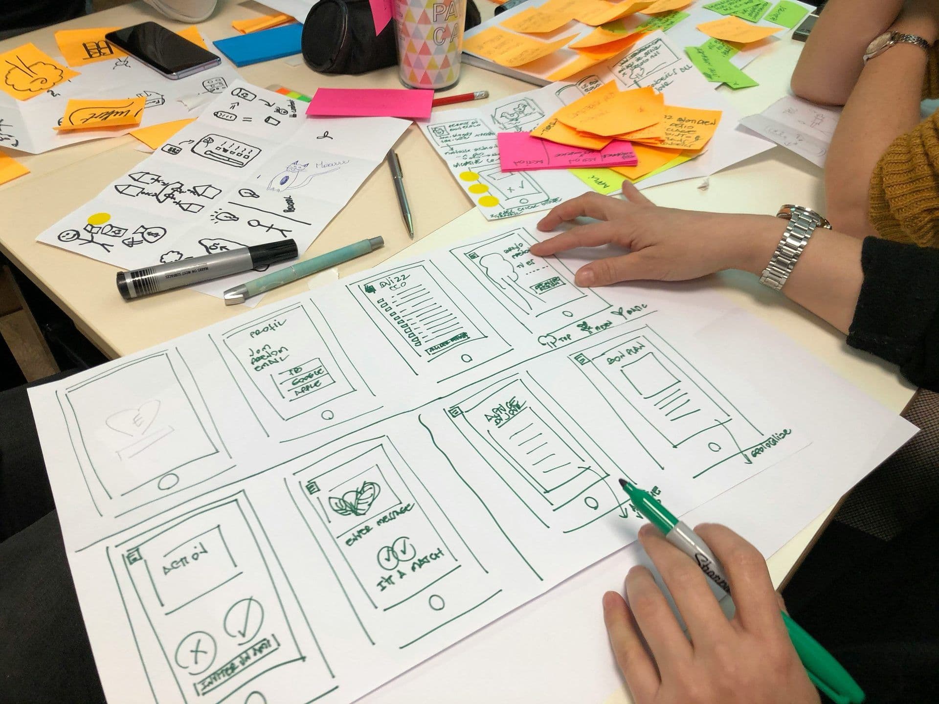 Hands sketching mobile app wireframes and ideas on a table during a collaborative design sprint workshop.