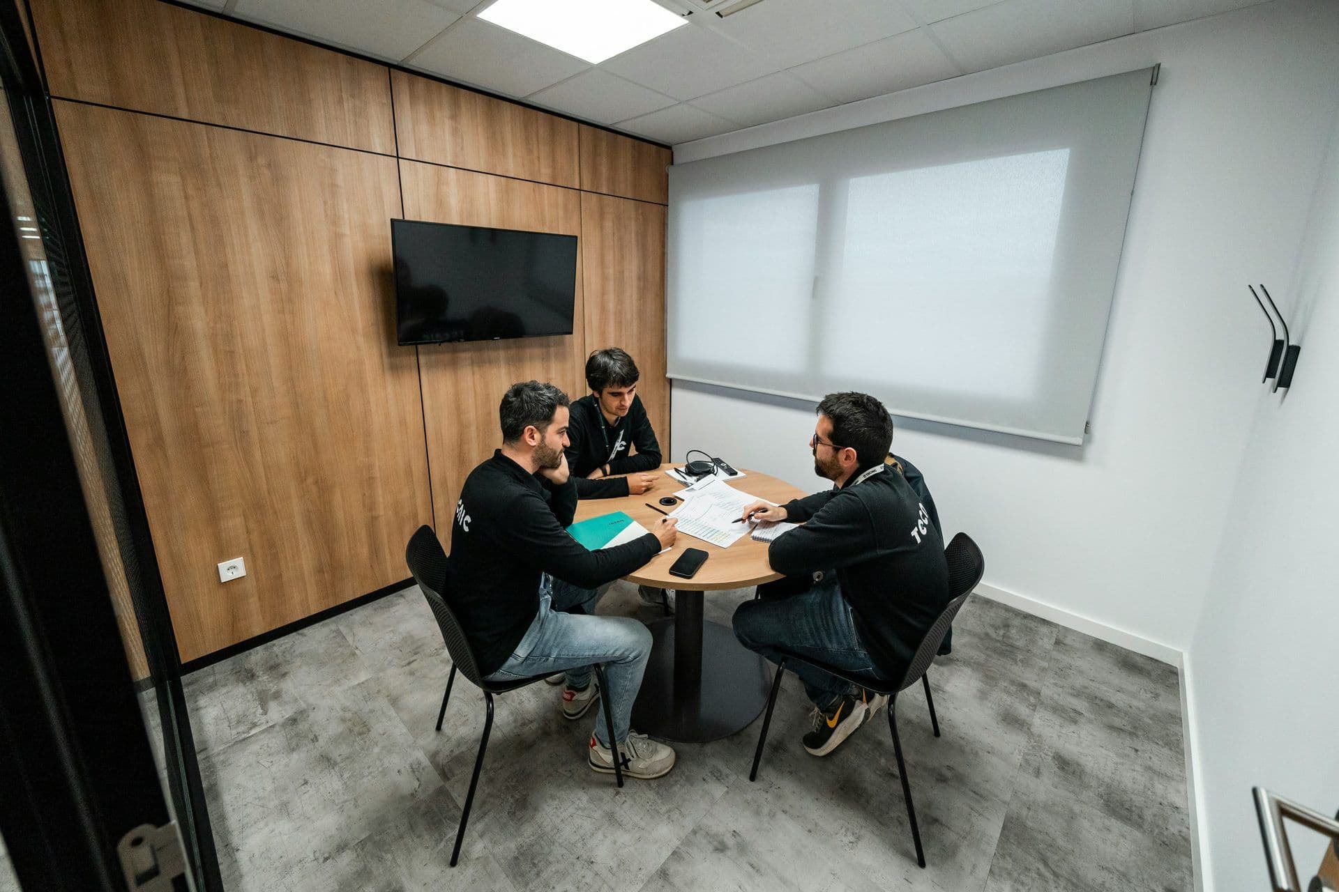 Three colleagues collaborate around a round table in a modern meeting room with a wall-mounted TV.