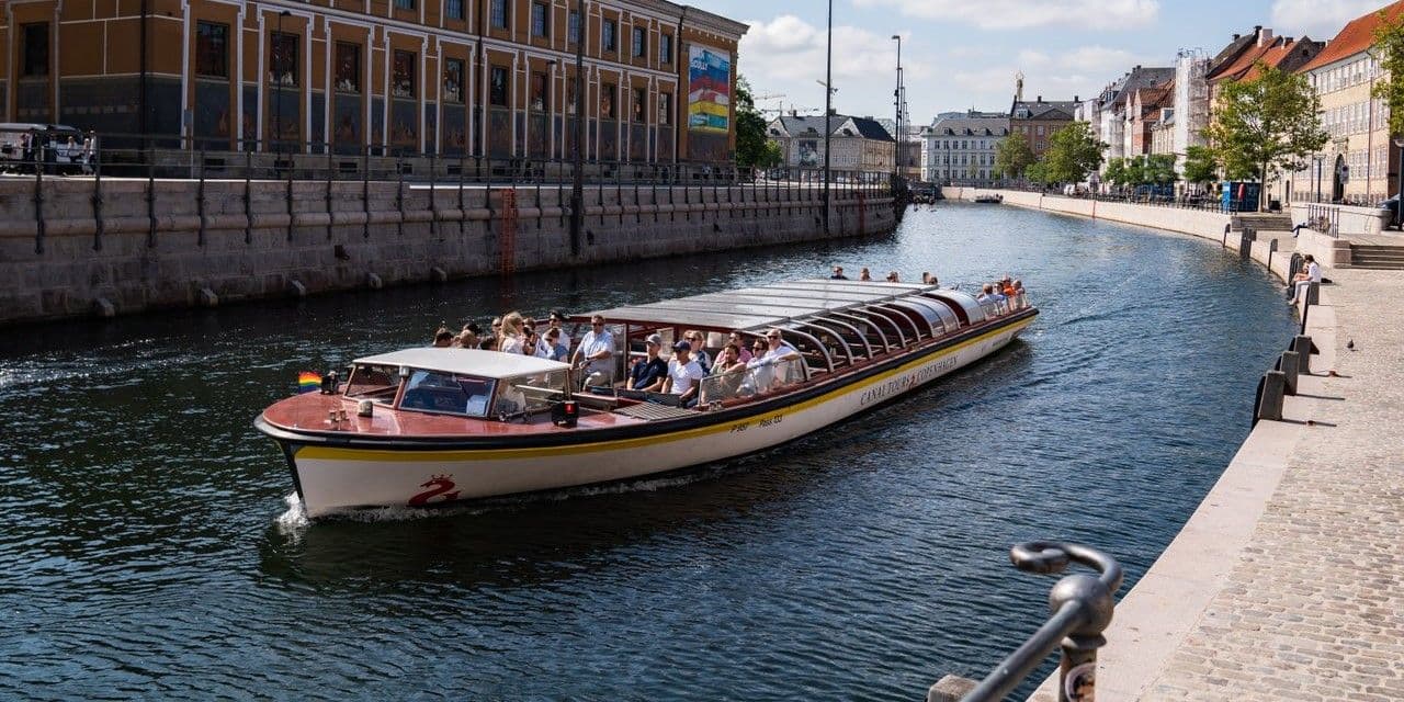 A large group on a canal boat tour in Copenhagen, passing historic buildings along the waterfront.