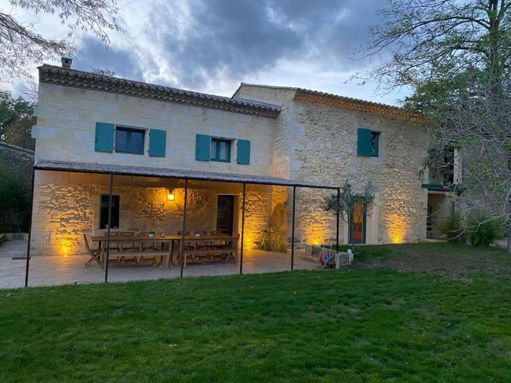 Stone villa with green shutters and illuminated outdoor patio featuring long wooden tables and benches.