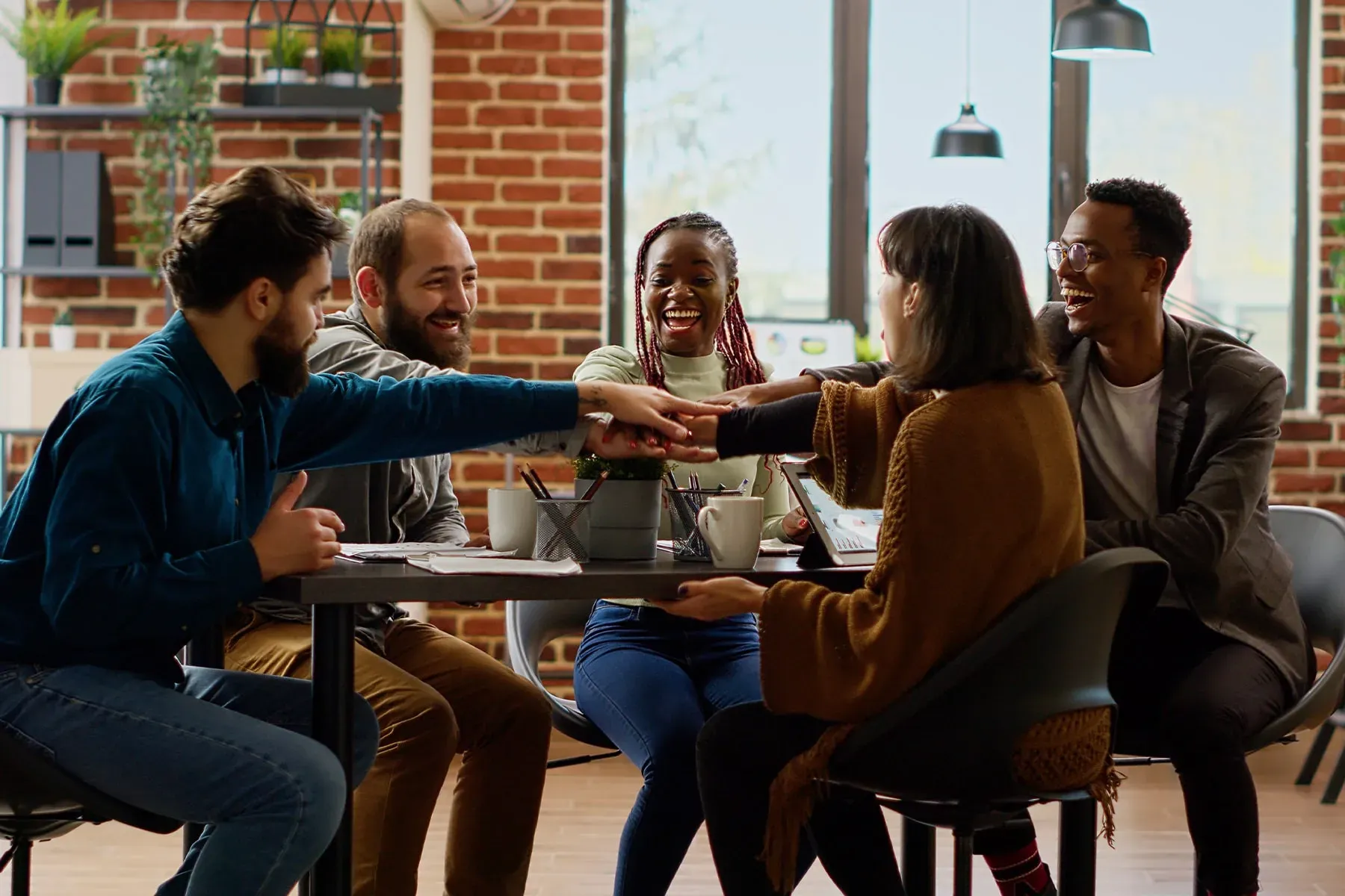 Five smiling colleagues stack hands at a table, demonstrating teamwork during a corporate workshop.