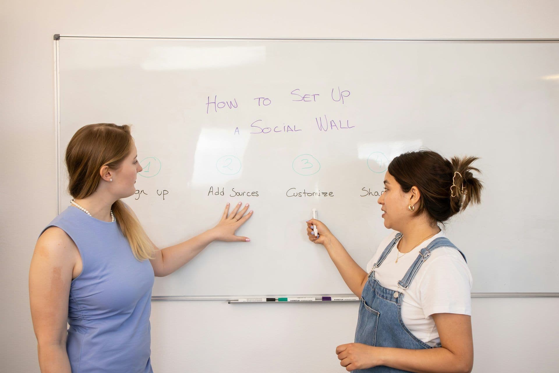 Two women present a 'How to Set Up A Social Wall' workshop on a whiteboard in a modern meeting room.