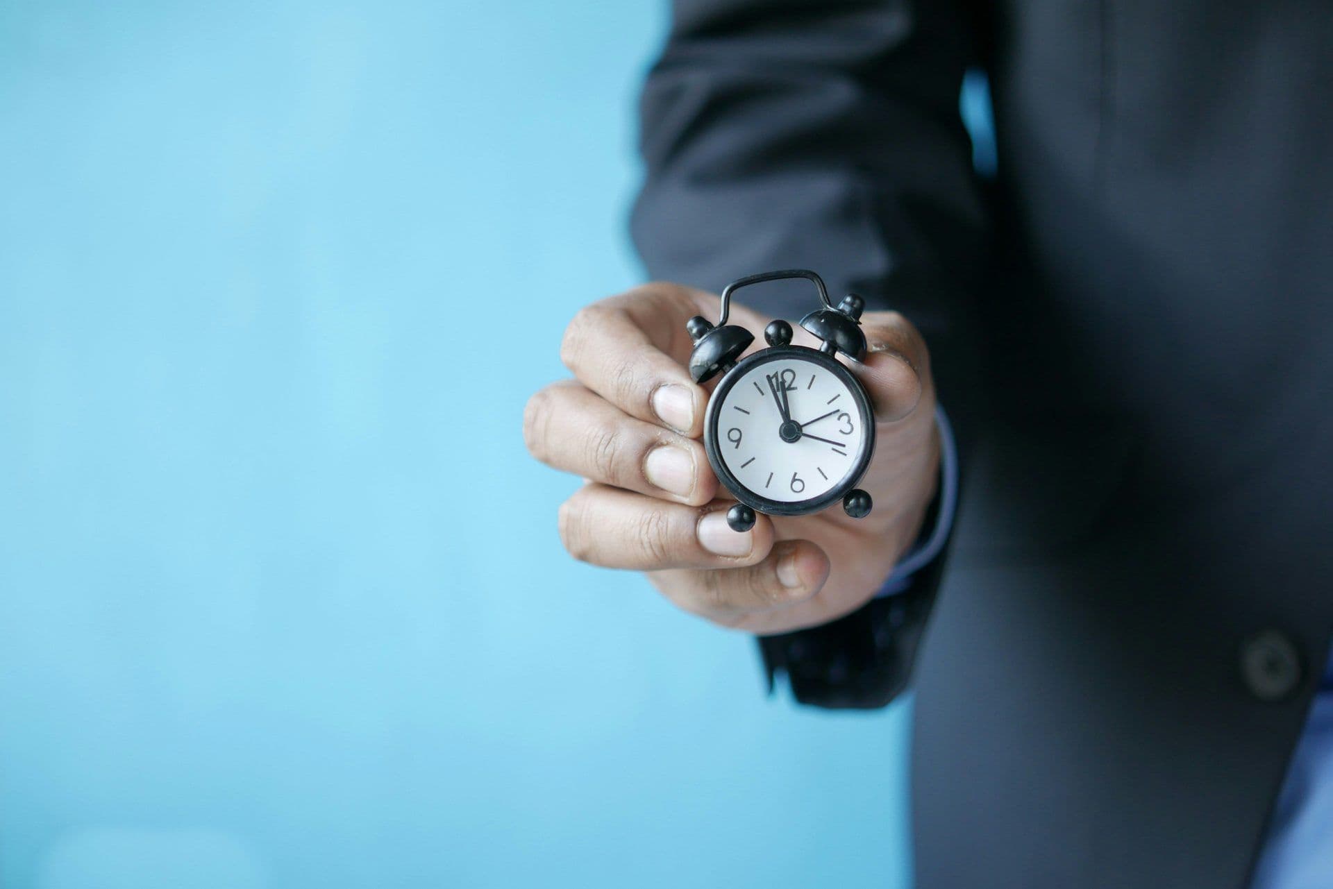 A hand in a dark suit holds a small black alarm clock, highlighting time management for corporate events.