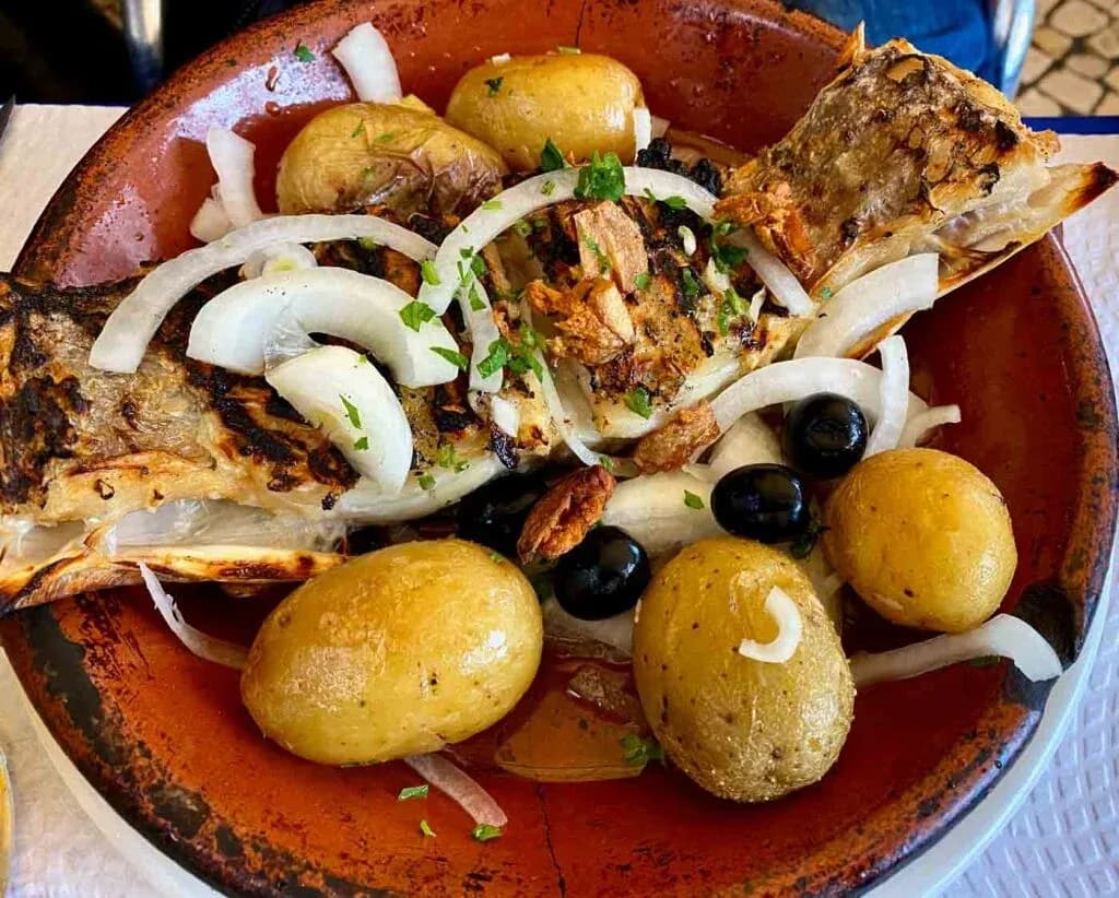 Grilled fish, roasted potatoes, onions, and olives in a terracotta dish, perfect for a corporate dinner.