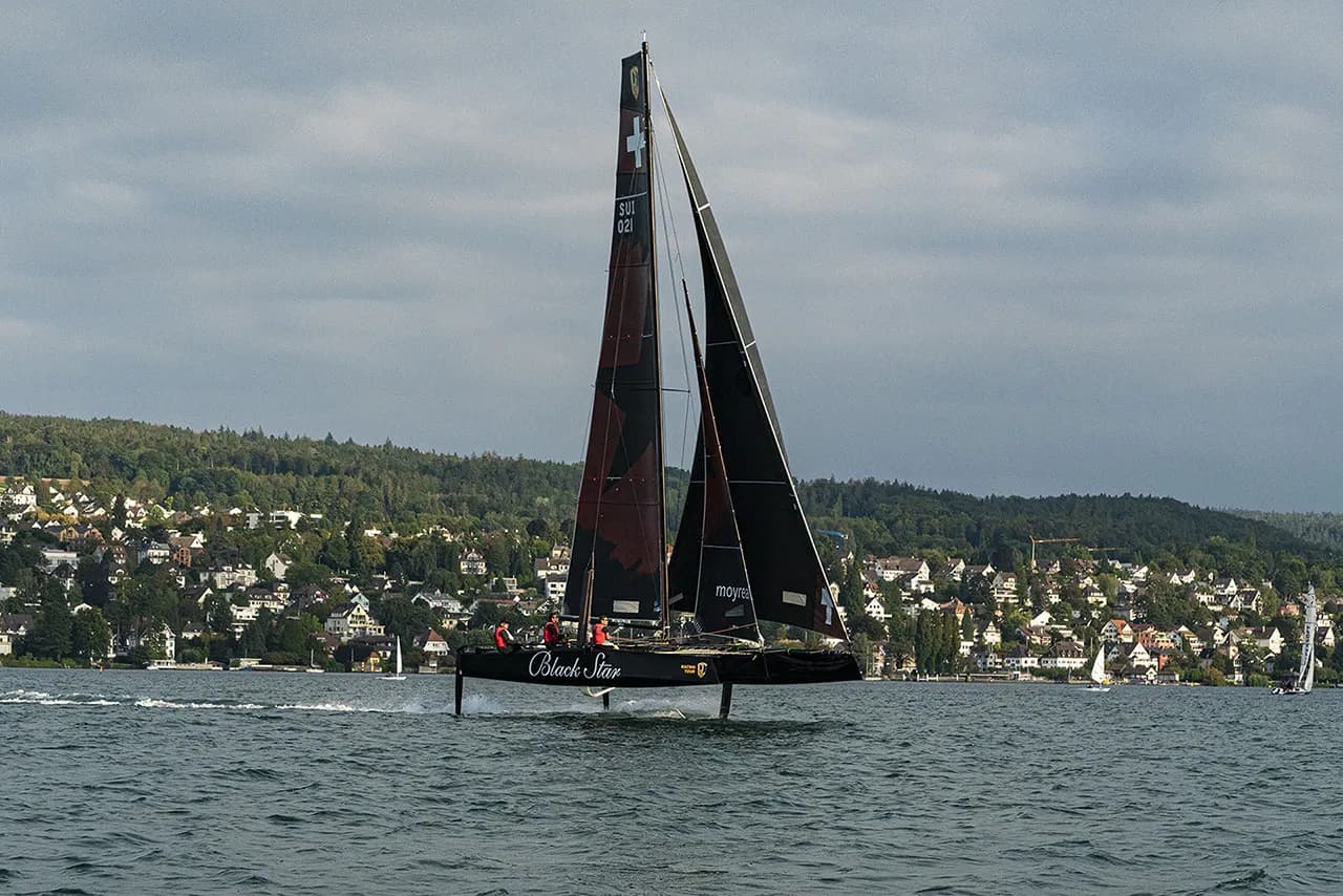 A black hydrofoil sailboat with a Swiss flag sails on Lake Zurich, past a tree-lined shore with houses.