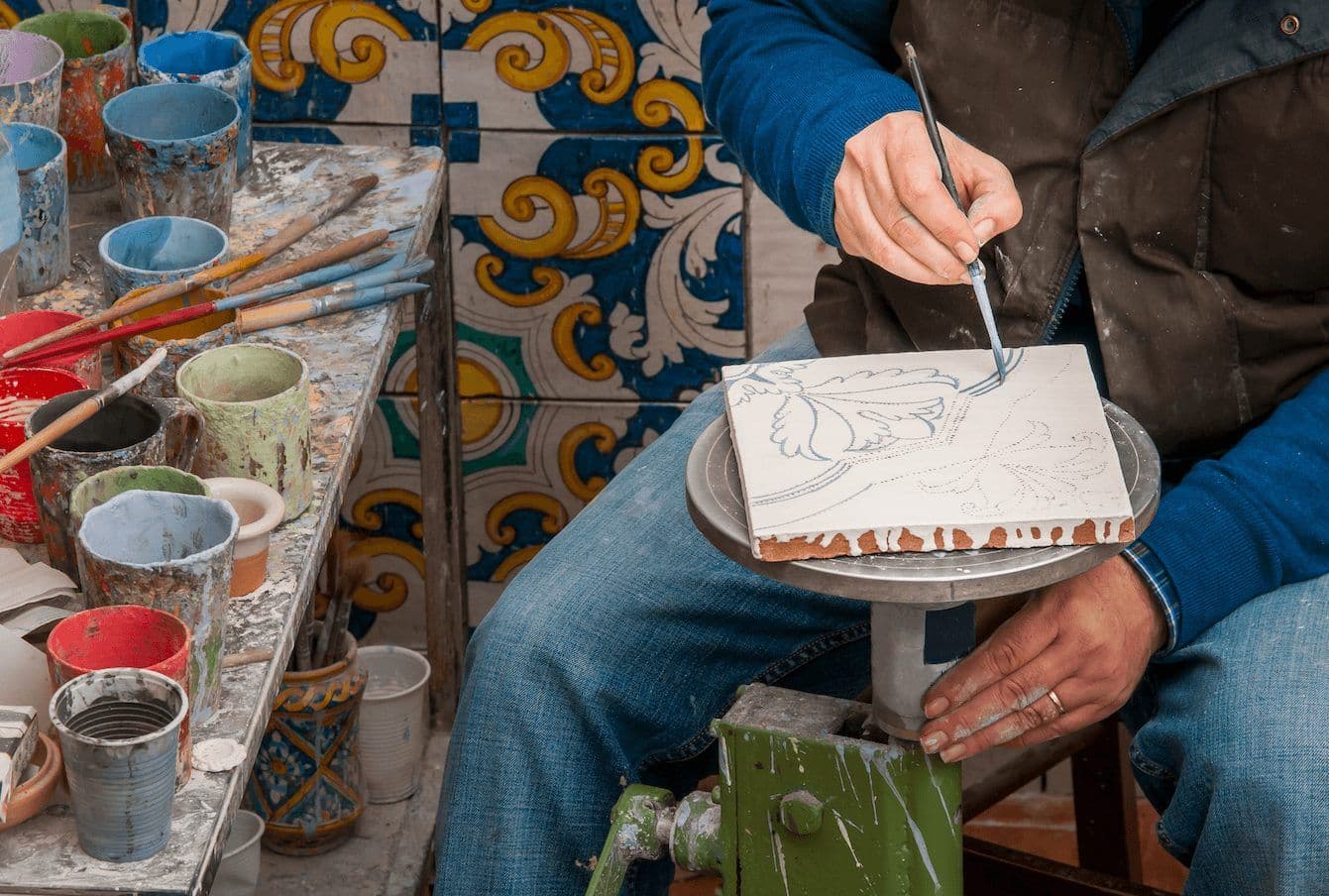 A person paints a decorative ceramic tile in an artisan workshop, surrounded by paint pots and brushes.