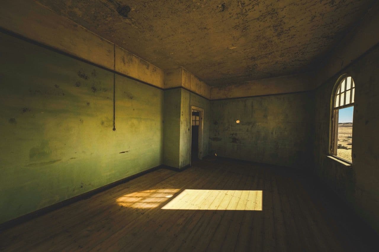 Historic room with peeling green walls, wooden floor, and arched window overlooking a desert.