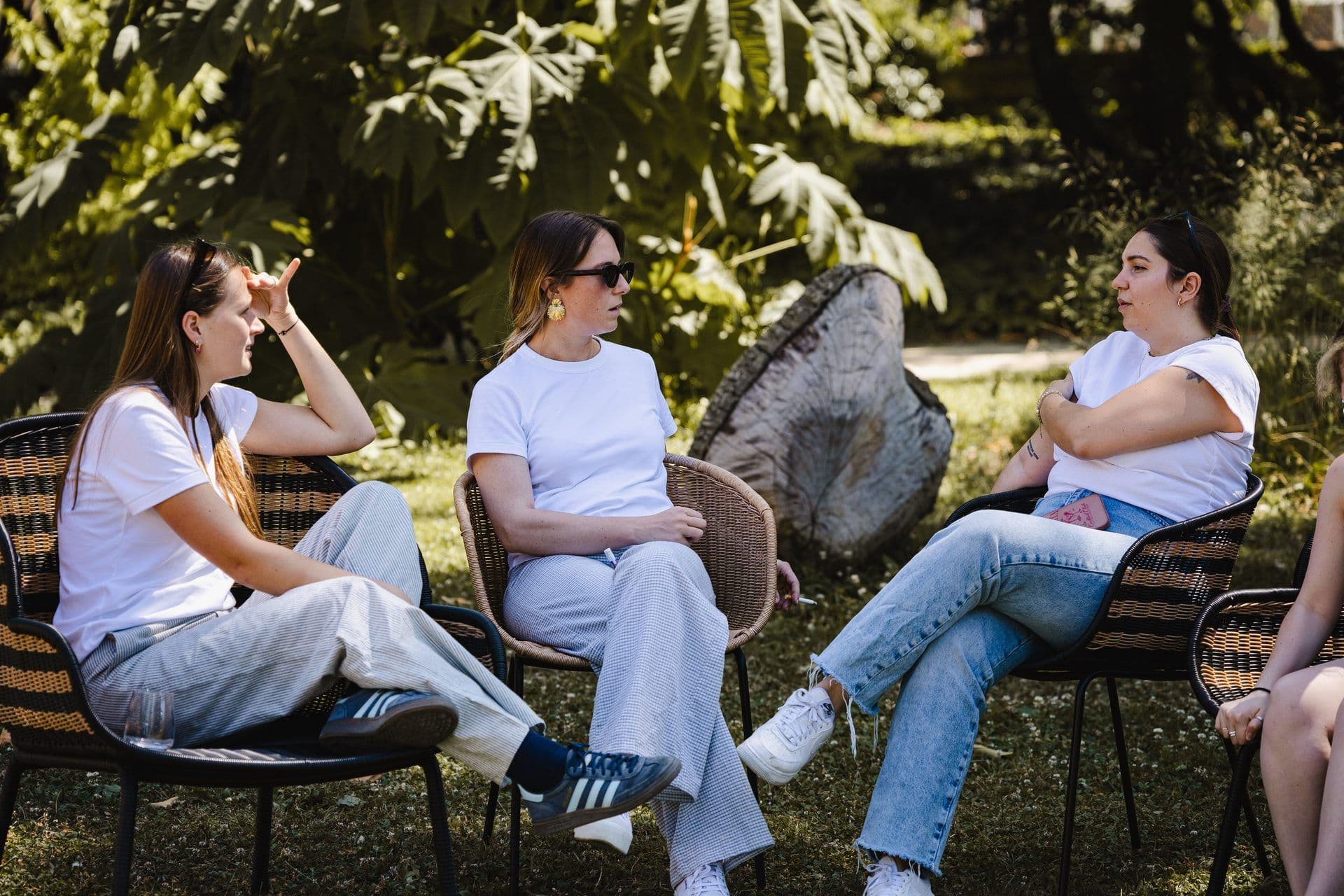 Three women in casual attire discuss in outdoor wicker chairs at a sunny garden offsite venue.