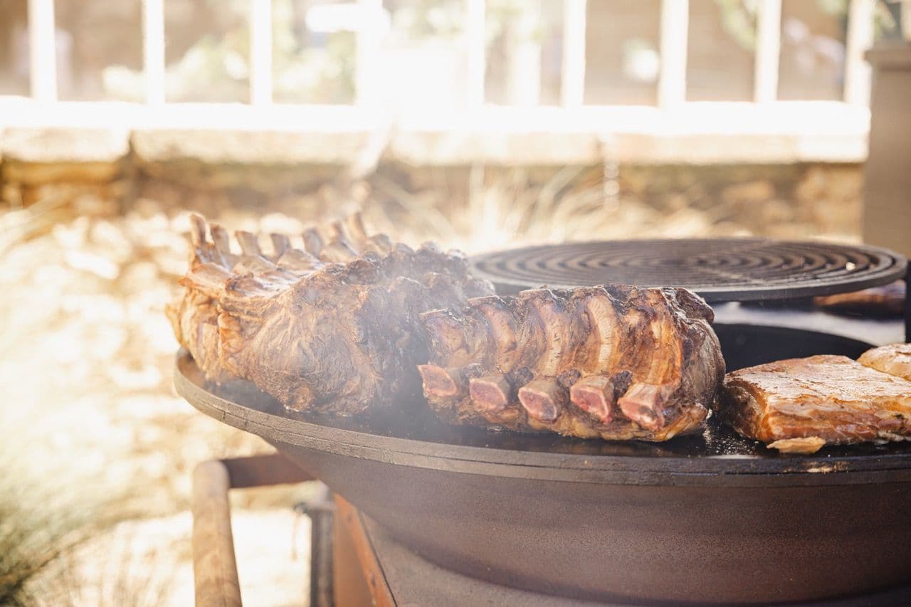 Large cuts of beef ribs and pork belly sizzle on an outdoor plancha grill for corporate catering.