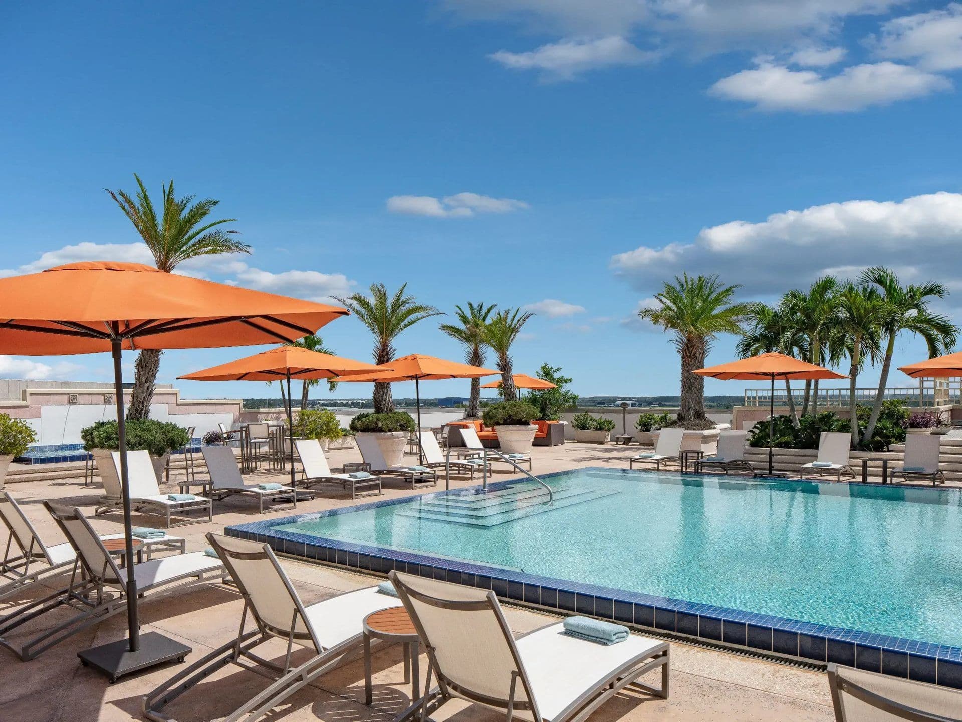 Outdoor rooftop pool deck in Orlando with lounge seating, orange umbrellas, and lush palm trees.