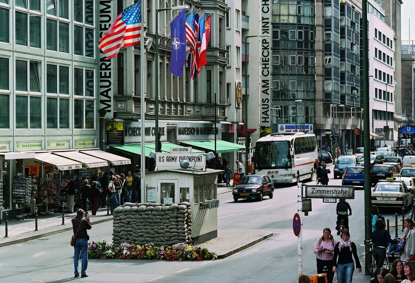 A busy street scene at Checkpoint Charlie in Berlin, featuring a guardhouse, flags, and museum.