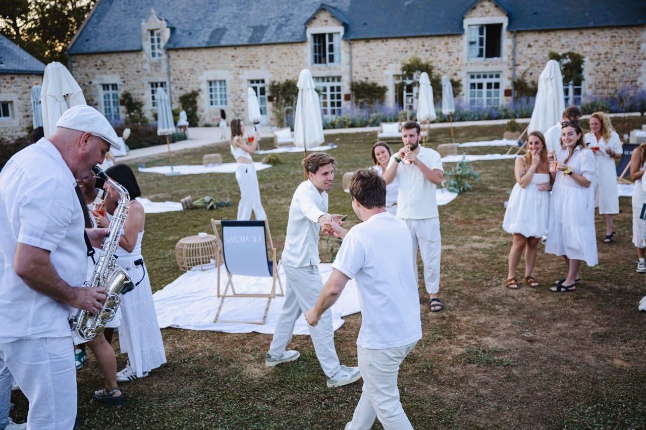 People in white enjoy an outdoor corporate cocktail reception with live saxophone music at a historic venue.