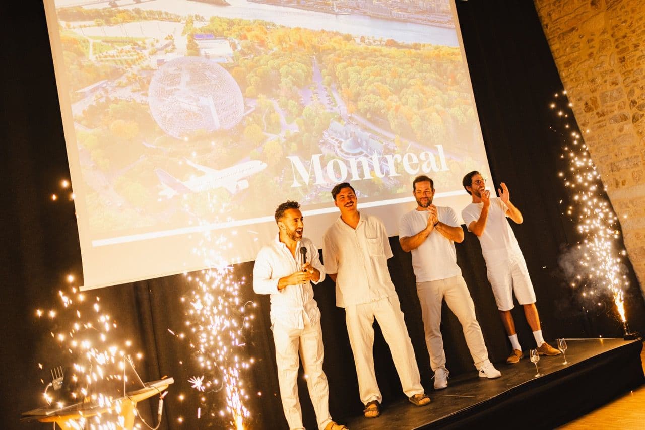 Four men on stage at a corporate event, with indoor fireworks and a large screen showing Montreal.