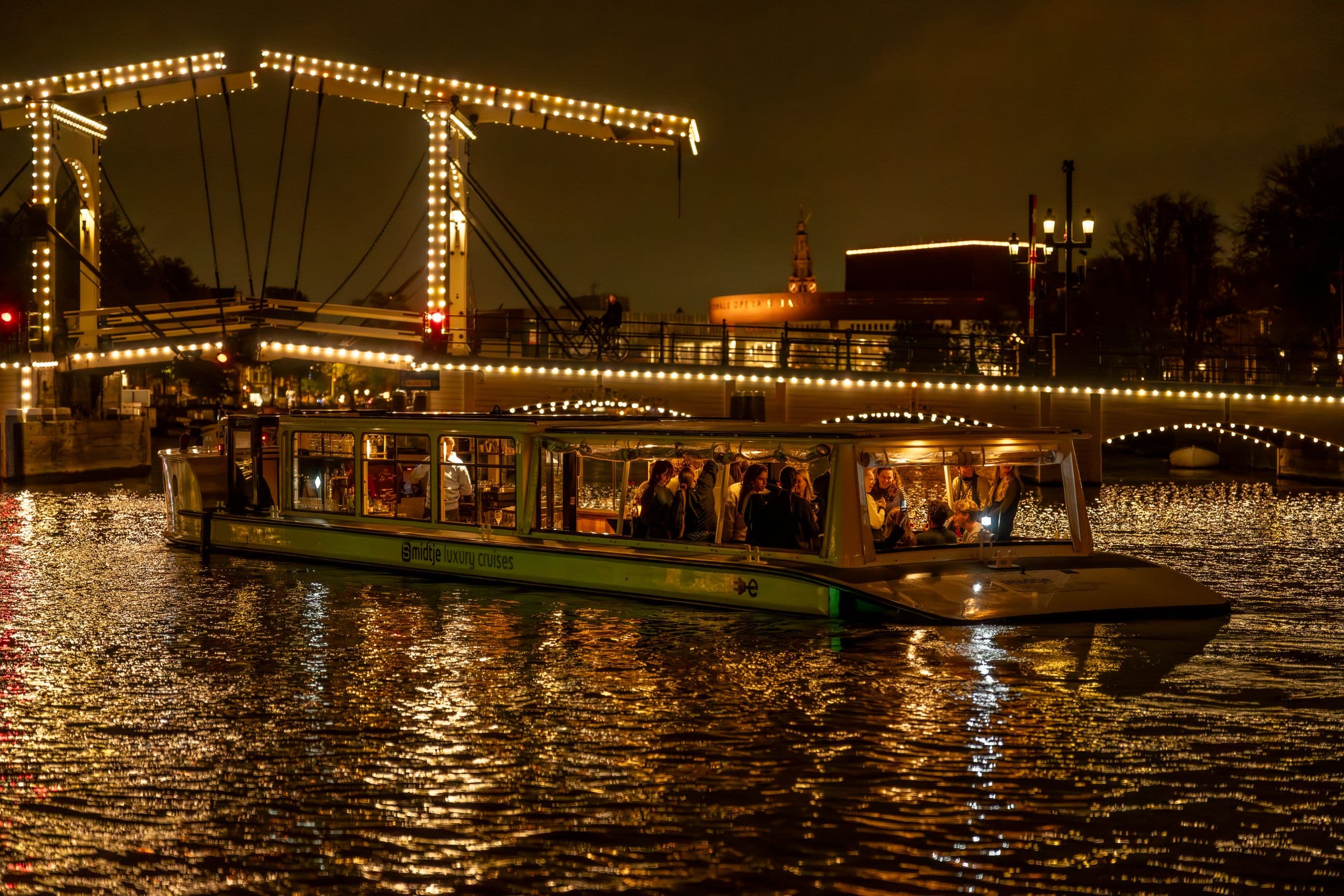 Illuminated canal boat with people enjoying a night cruise in Amsterdam, passing a lit drawbridge.