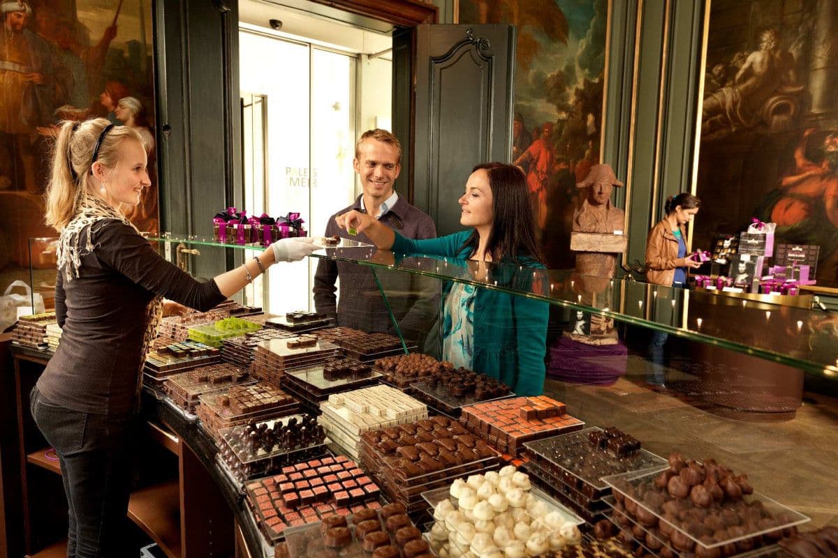 A sales assistant serves a couple sampling artisanal chocolates from a large display in a historic venue.