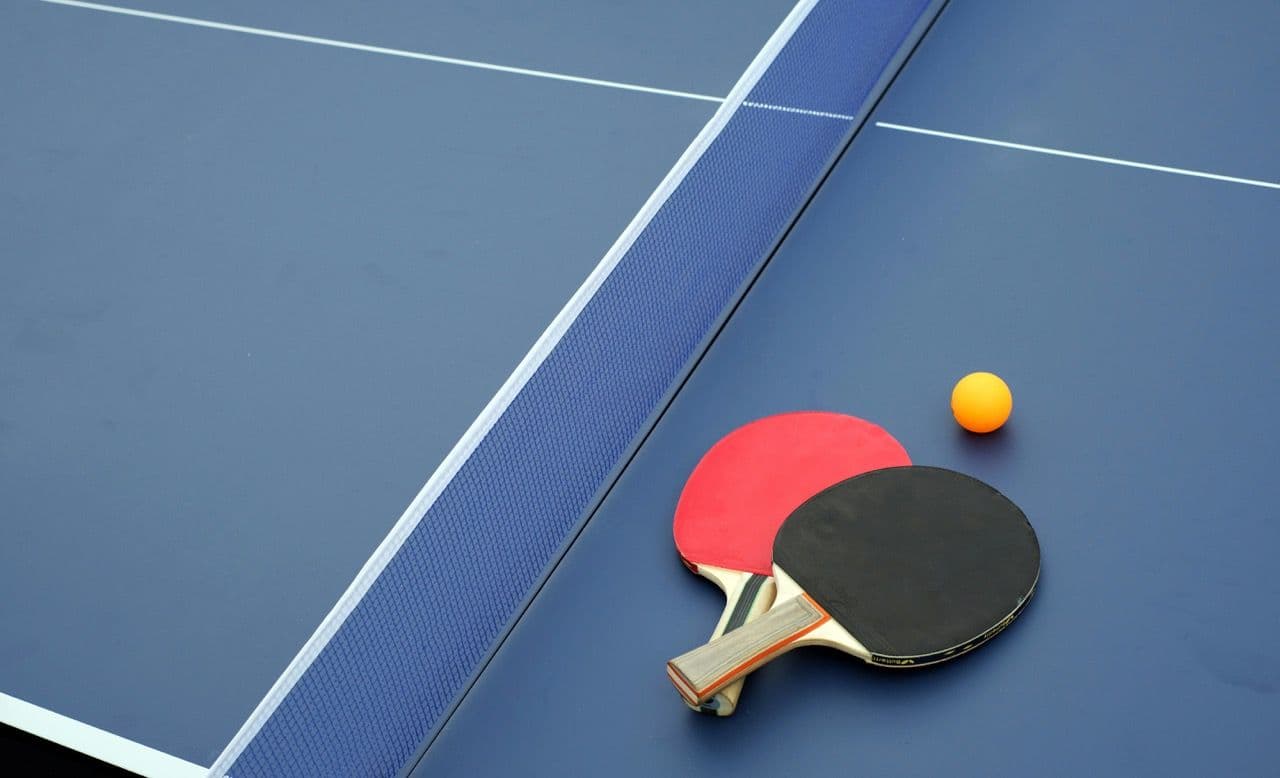 Two ping-pong paddles and an orange ball rest on a blue table with a net, suggesting a team building activity.