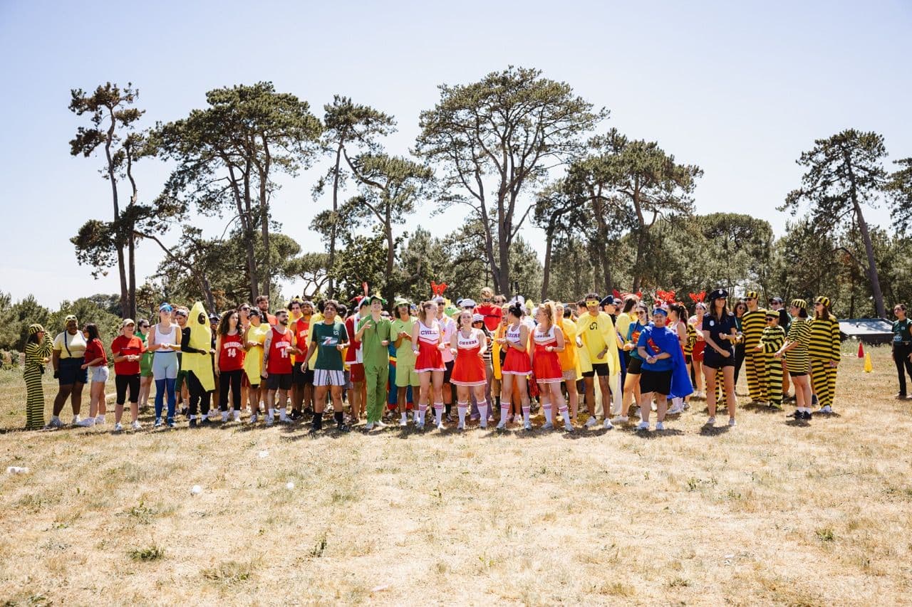 A large group of employees in various costumes stands in a grassy field during an outdoor team building