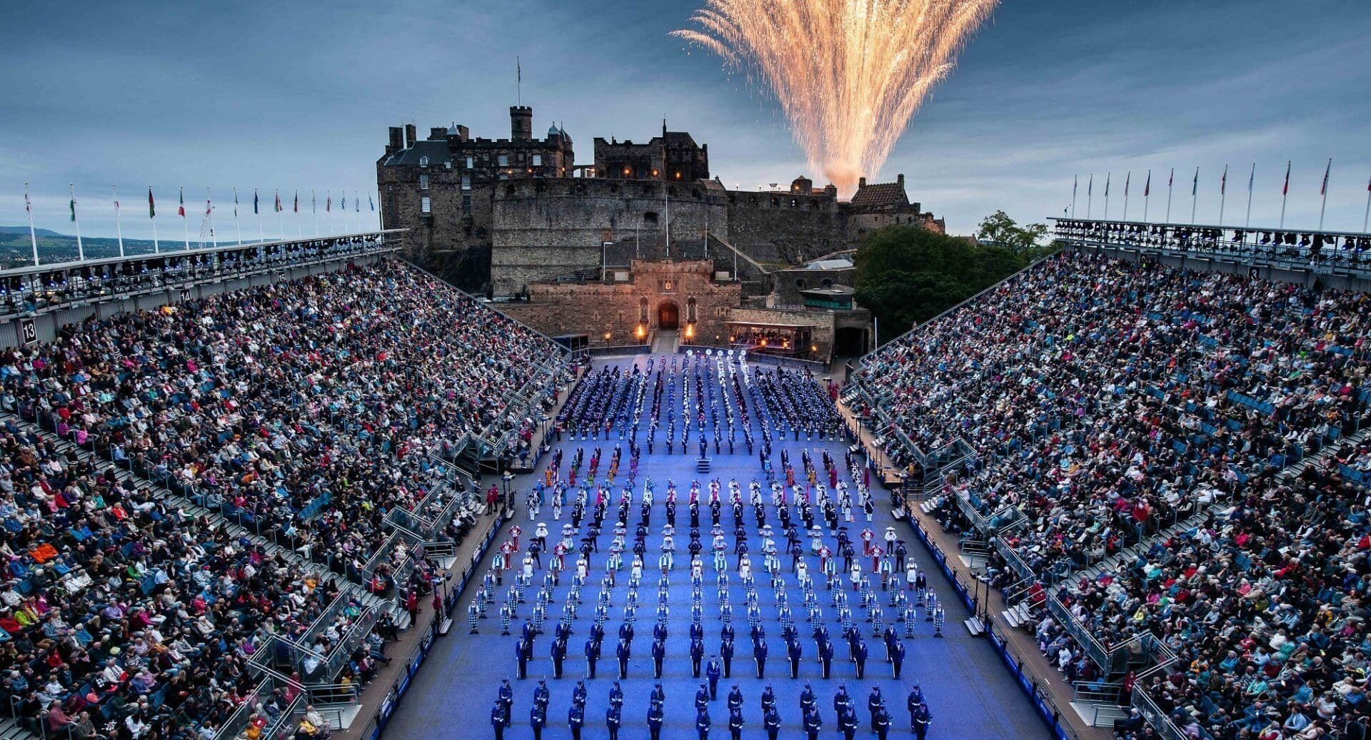 Fireworks illuminate Edinburgh Castle during a large outdoor corporate event with performers and a huge