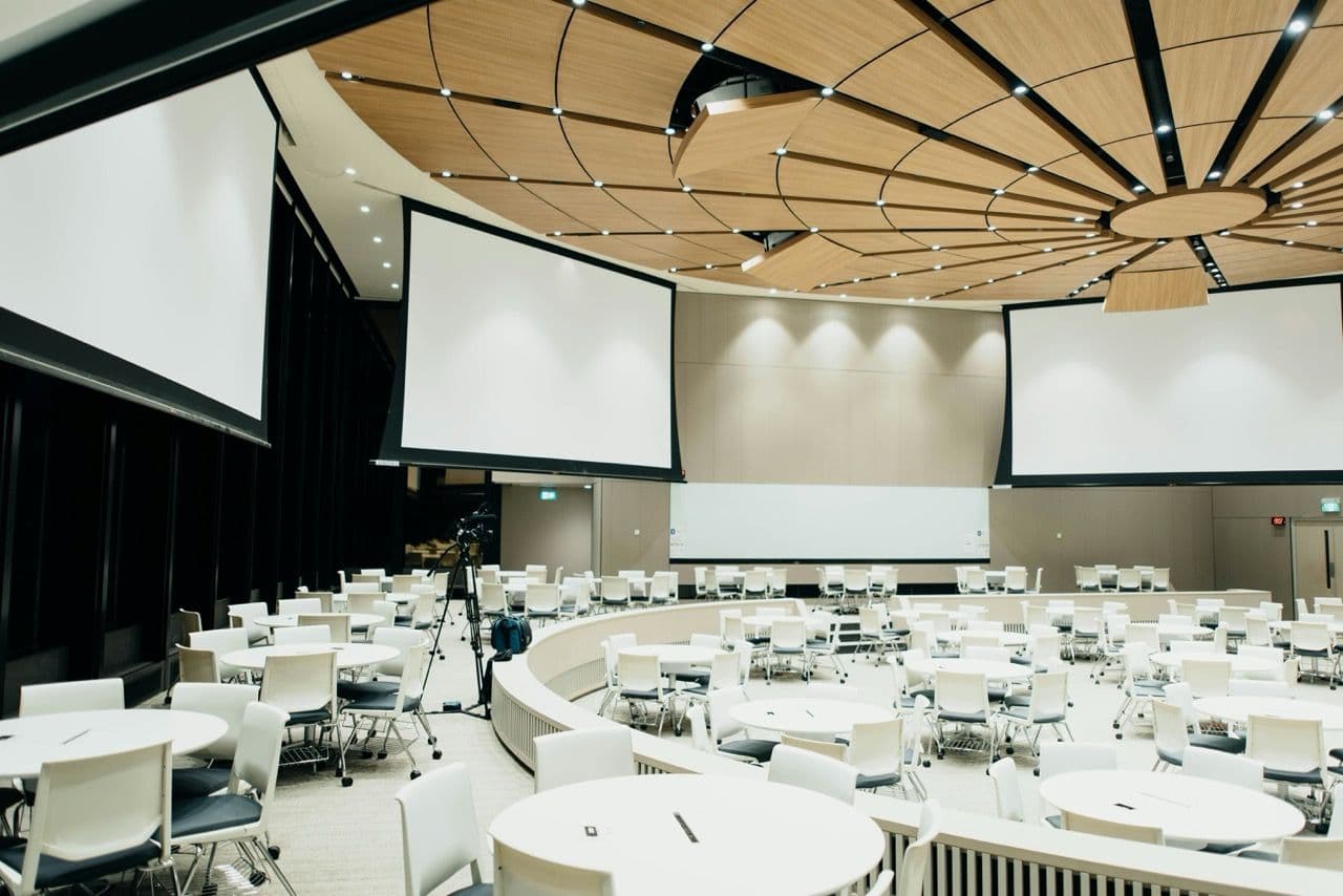 Spacious conference room set up with round tables, chairs, and large screens for a corporate seminar.