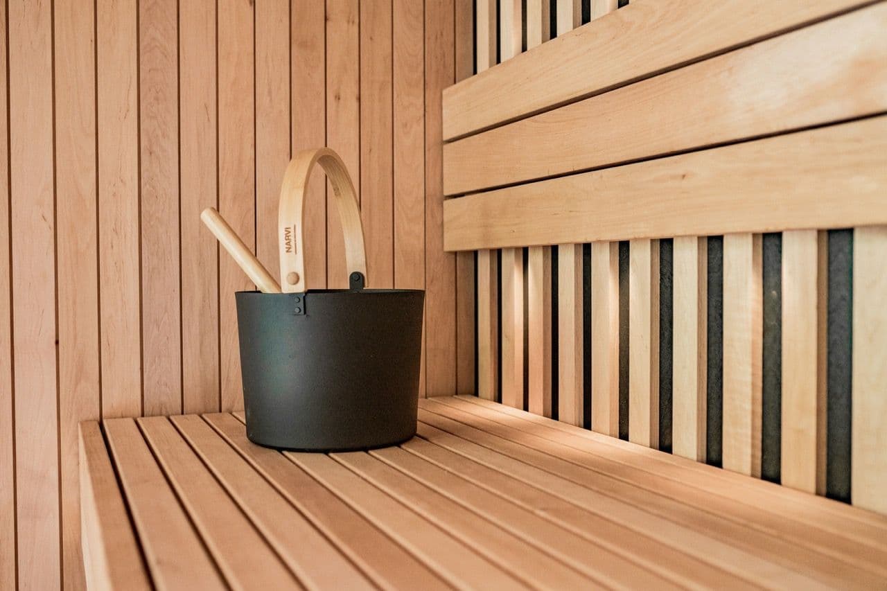 A modern wooden sauna interior featuring a black bucket and ladle on a slatted bench.