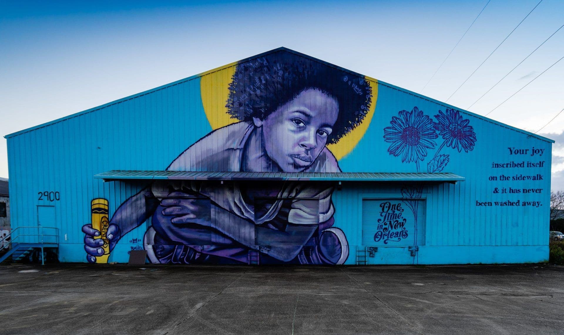 Vibrant mural of a child holding a yellow crayon on a blue building in New Orleans under a clear sky.