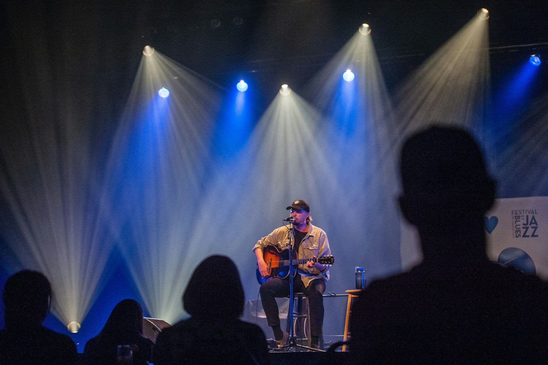 A male musician performs live with an acoustic guitar on a stage lit by blue and white spotlights.