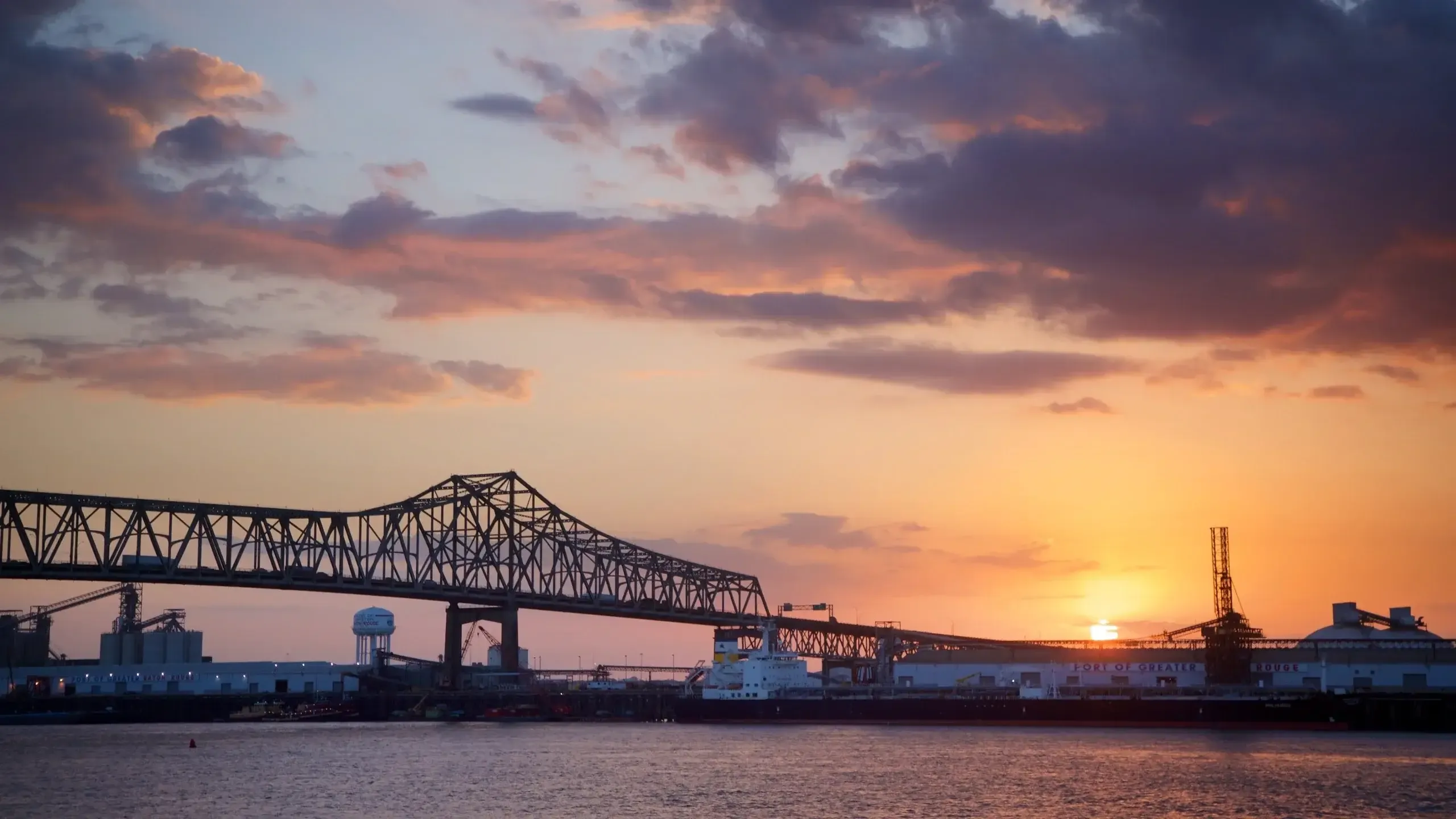 Sunset over the Mississippi River in Baton Rouge, Louisiana, with a truss bridge and industrial port.