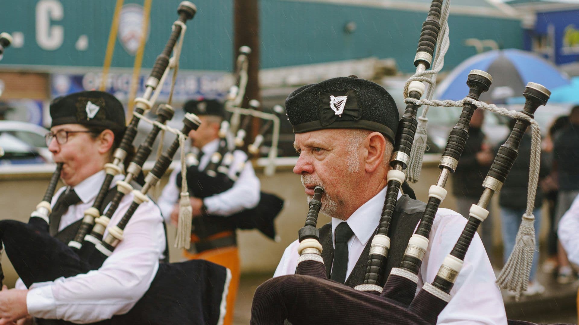Two bagpipers in traditional Irish uniform perform outdoors in the rain