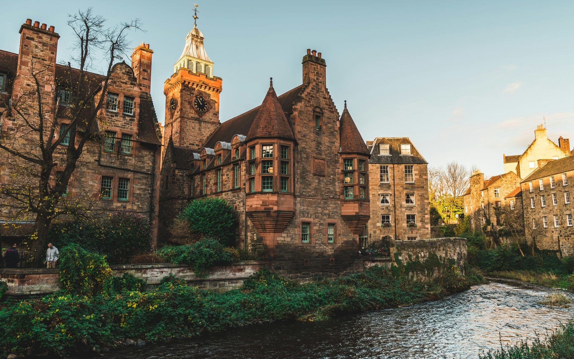 Historic stone building with a prominent clock tower and green windows overlooking a river in Edinburgh