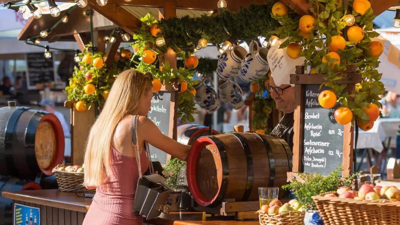 Outdoor festival stall in Frankfurt, Germany, with a woman ordering apple wine from a vendor.