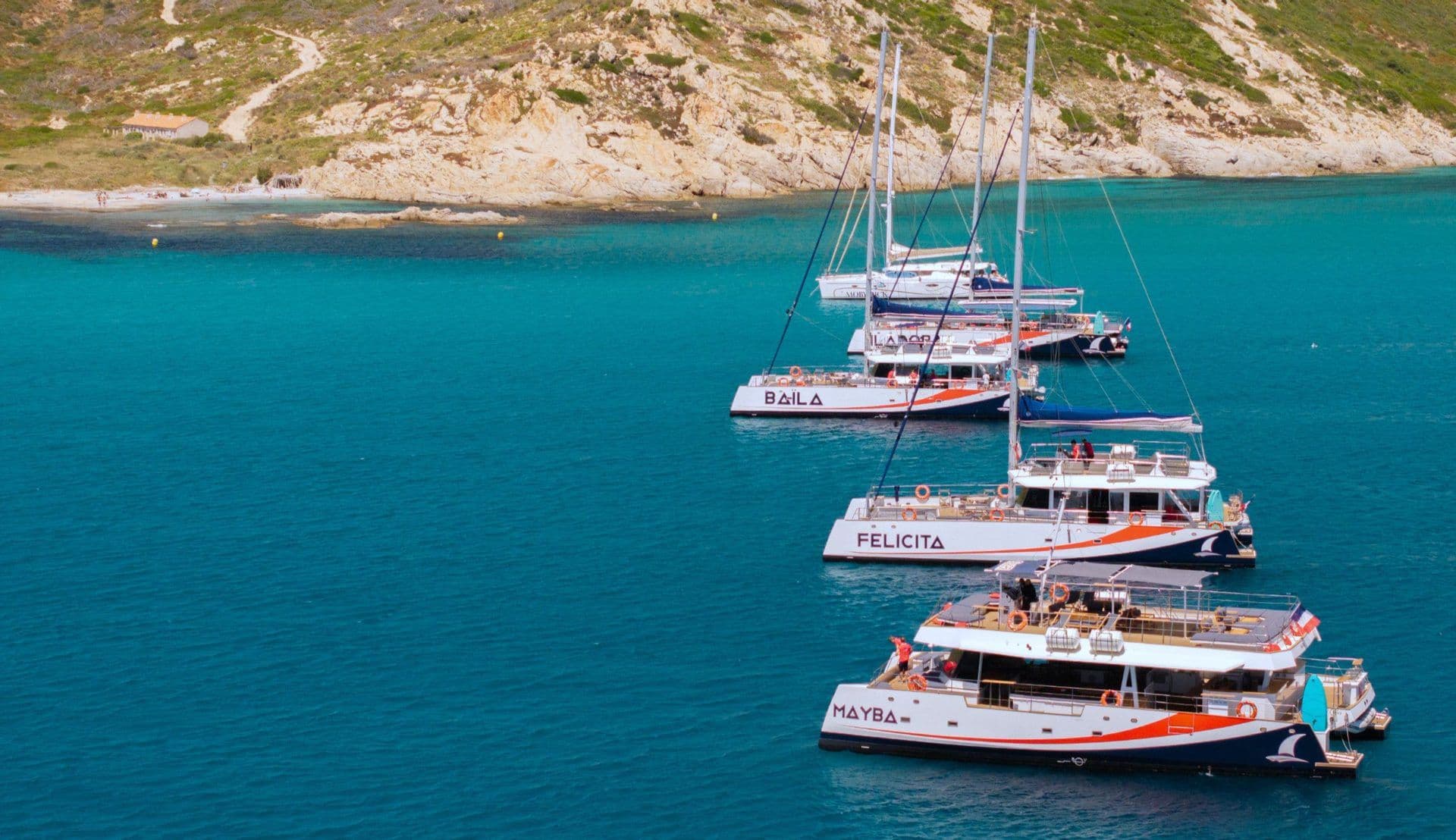 Several catamarans anchored in turquoise water near a rocky coastline, ideal for a corporate sailing event.
