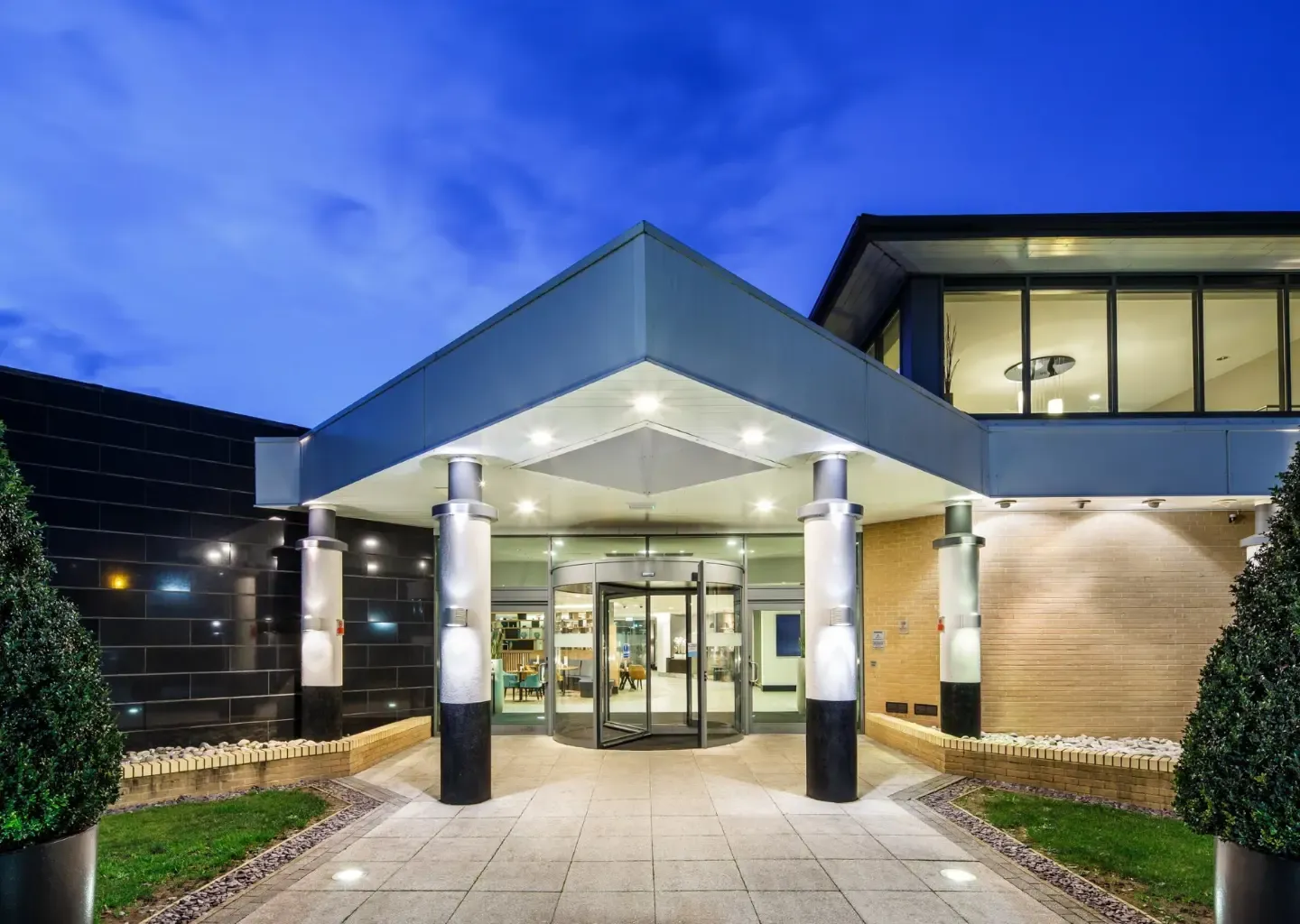 Illuminated modern corporate event venue entrance with a revolving door and covered portico at dusk.
