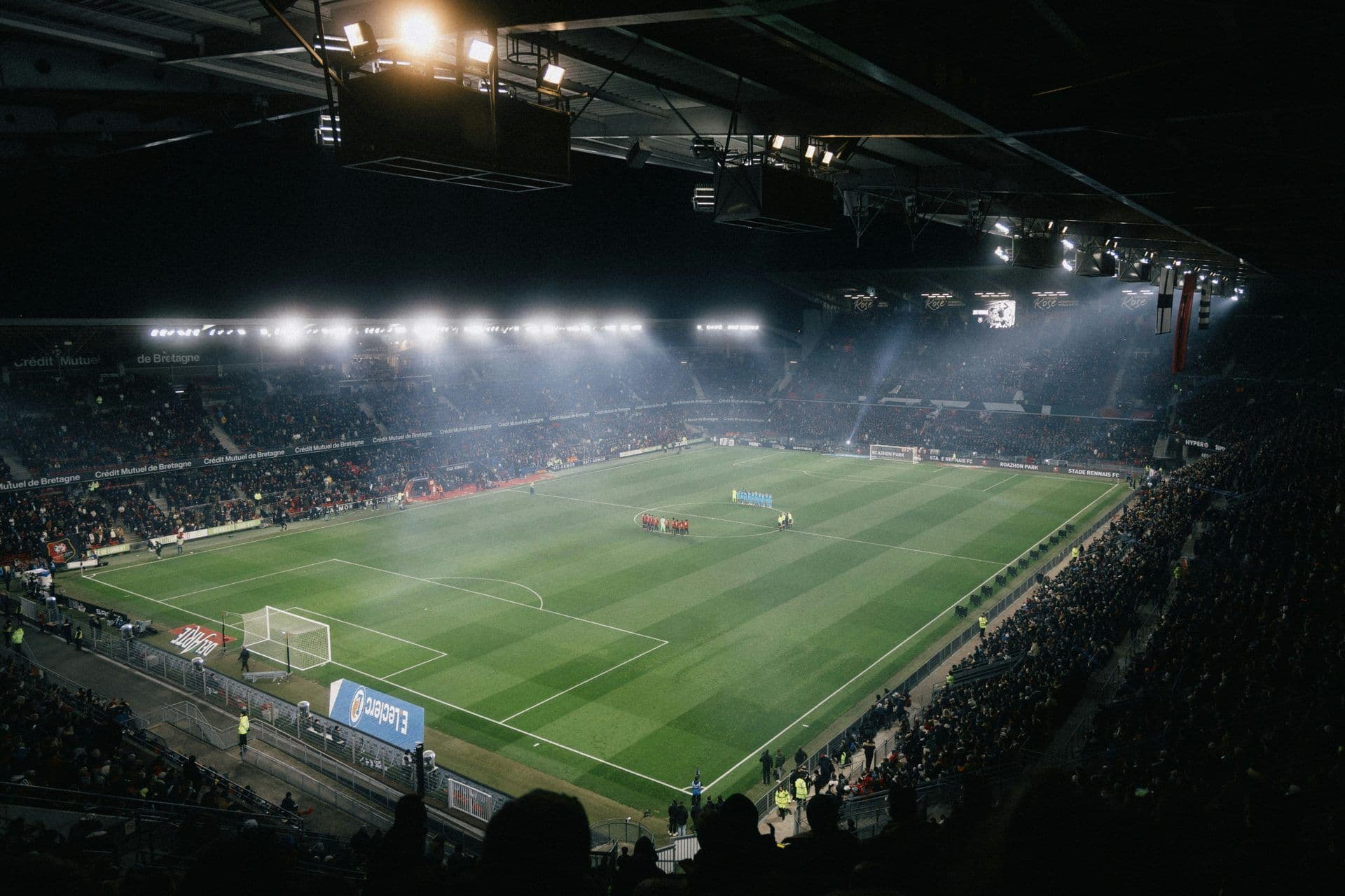 An illuminated football stadium in Rennes, France, hosting a night game with players on the pitch and a large