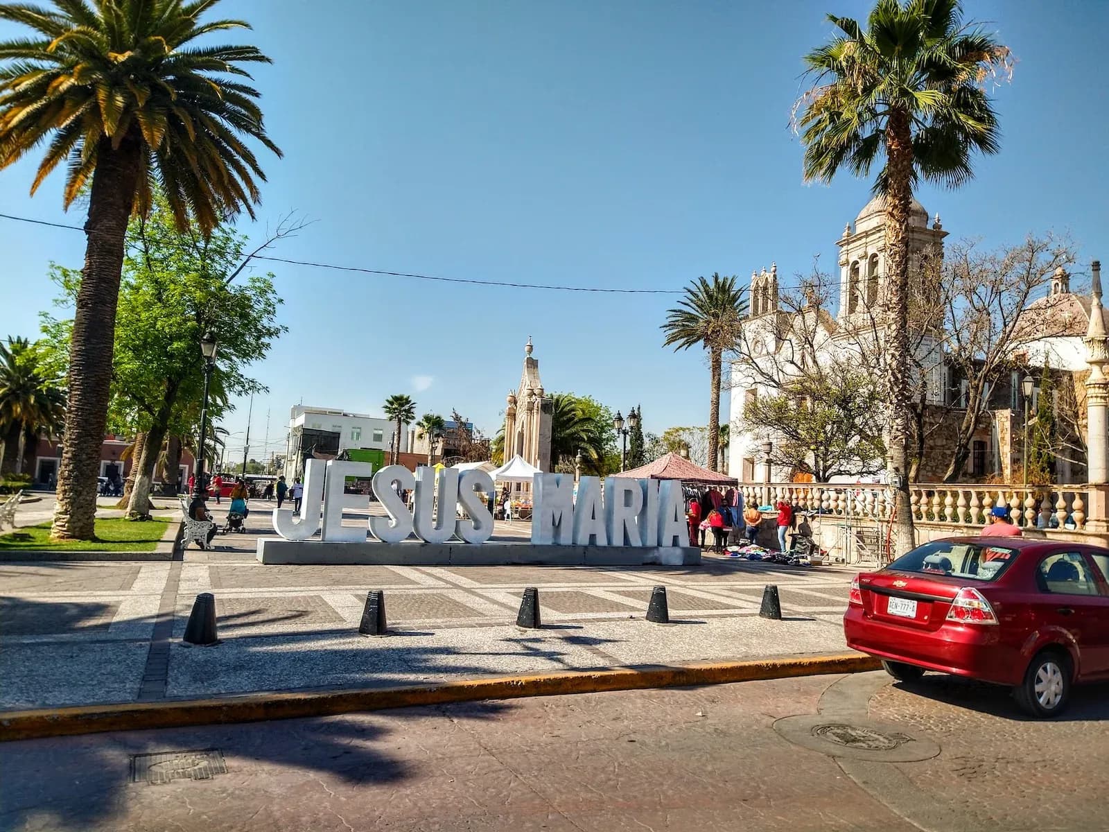 Large 'JESUS MARIA' sign in a sunny town square with palm trees and a historic church in Mexico.