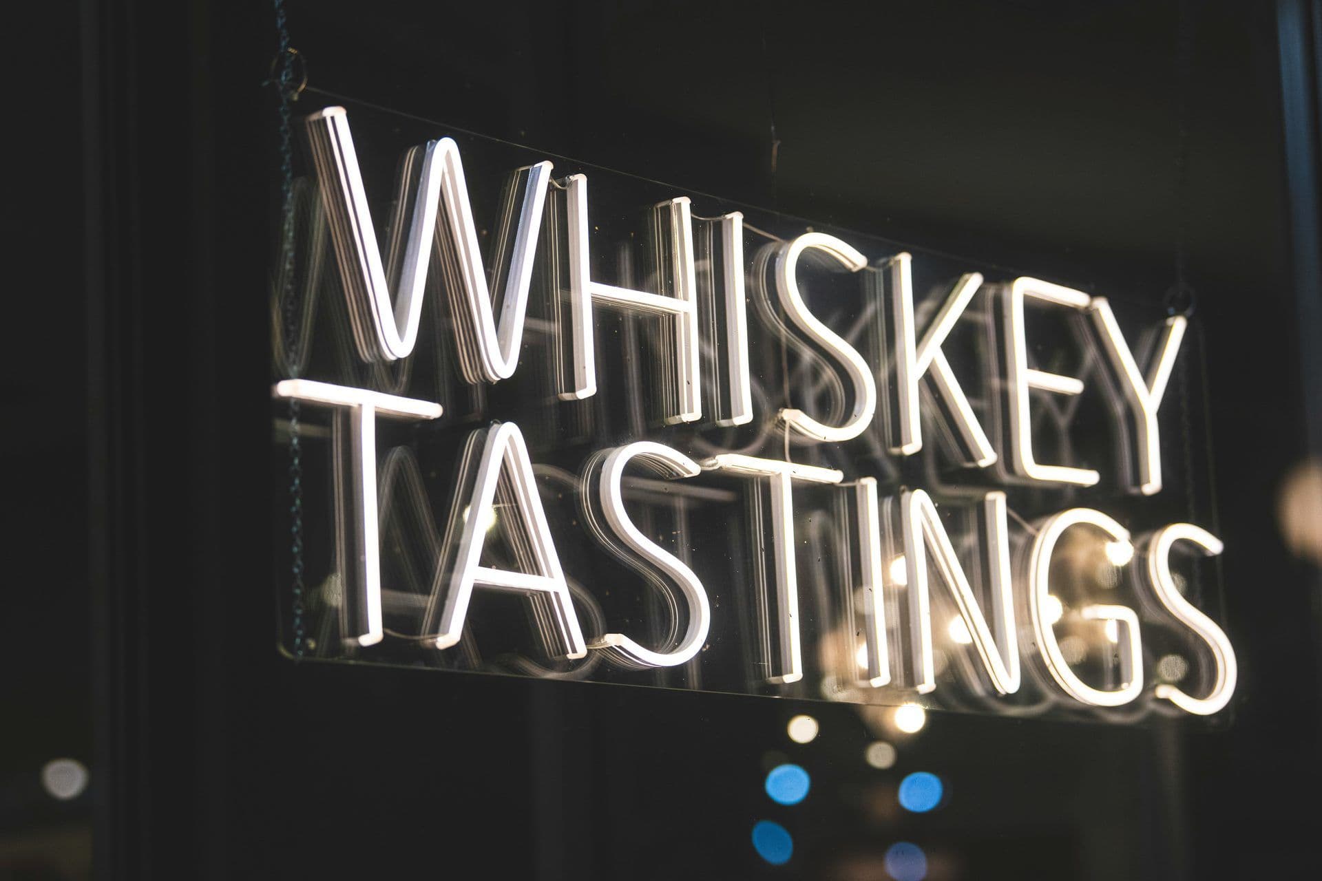 A bright white neon sign advertises 'WHISKEY TASTINGS' in a dark window at night.