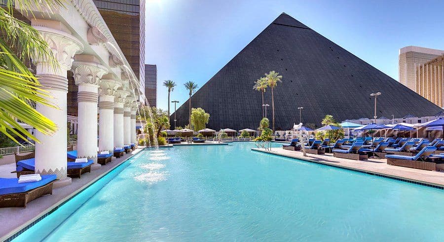 Large outdoor pool area with blue lounge chairs, palm trees, and the Luxor pyramid in Las Vegas.