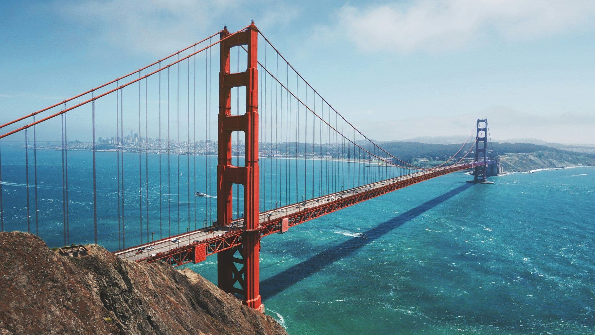 Panoramic view of the Golden Gate Bridge spanning the San Francisco Bay with city skyline and cliffs.
