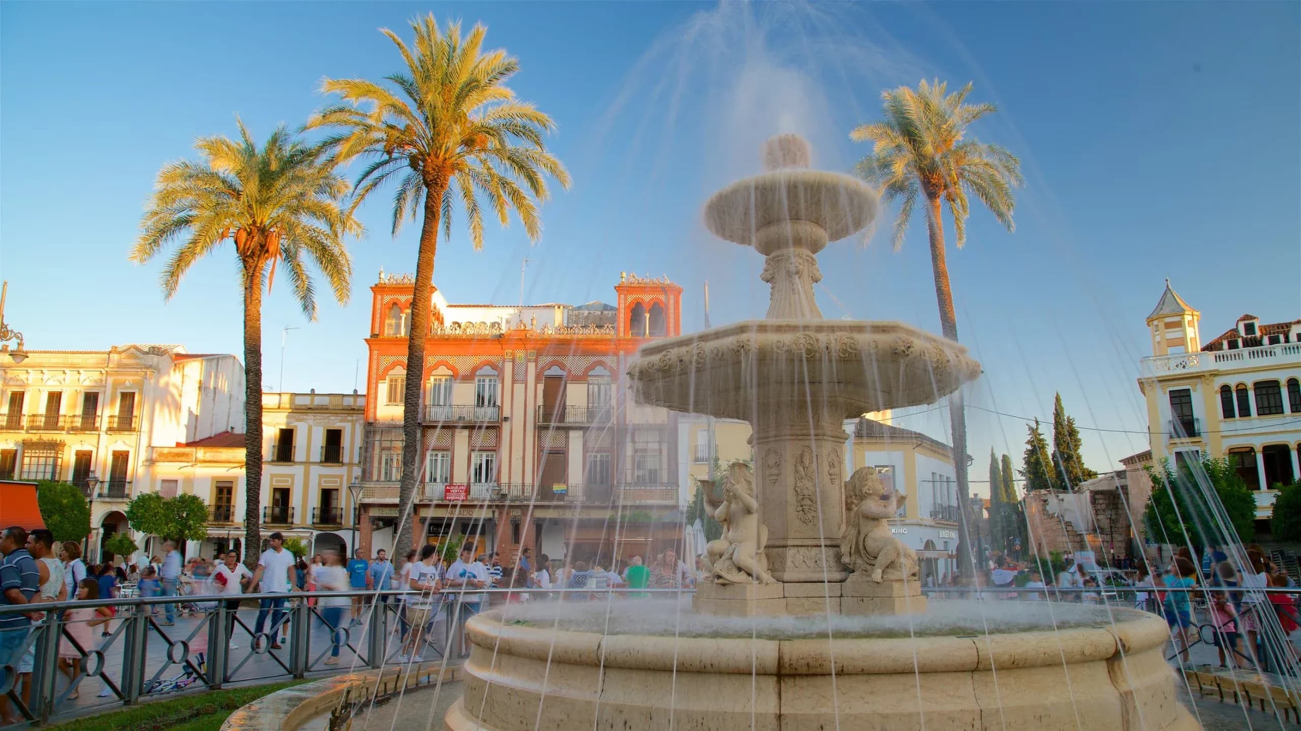 An ornate stone fountain with palm trees stands in a sunny historic square in Merida, Spain.