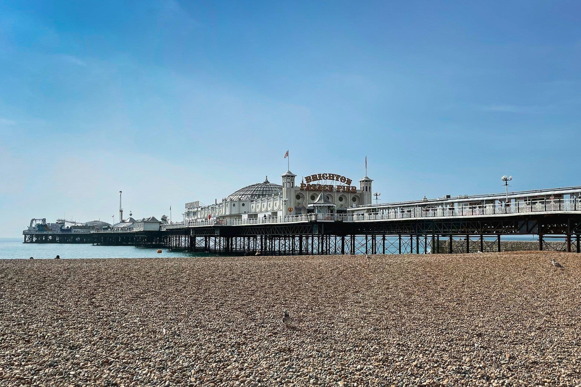 Brighton Palace Pier extends over the sea from a pebble beach under a clear blue sky in Brighton, UK.