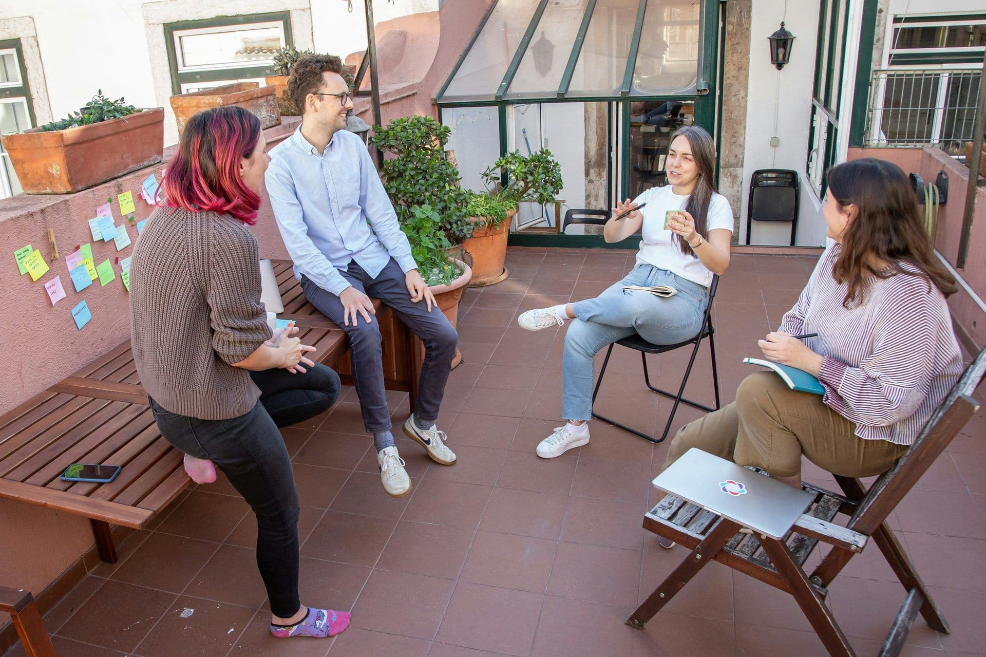Four colleagues hold a casual team offsite meeting on an outdoor terrace with sticky notes on the wall.