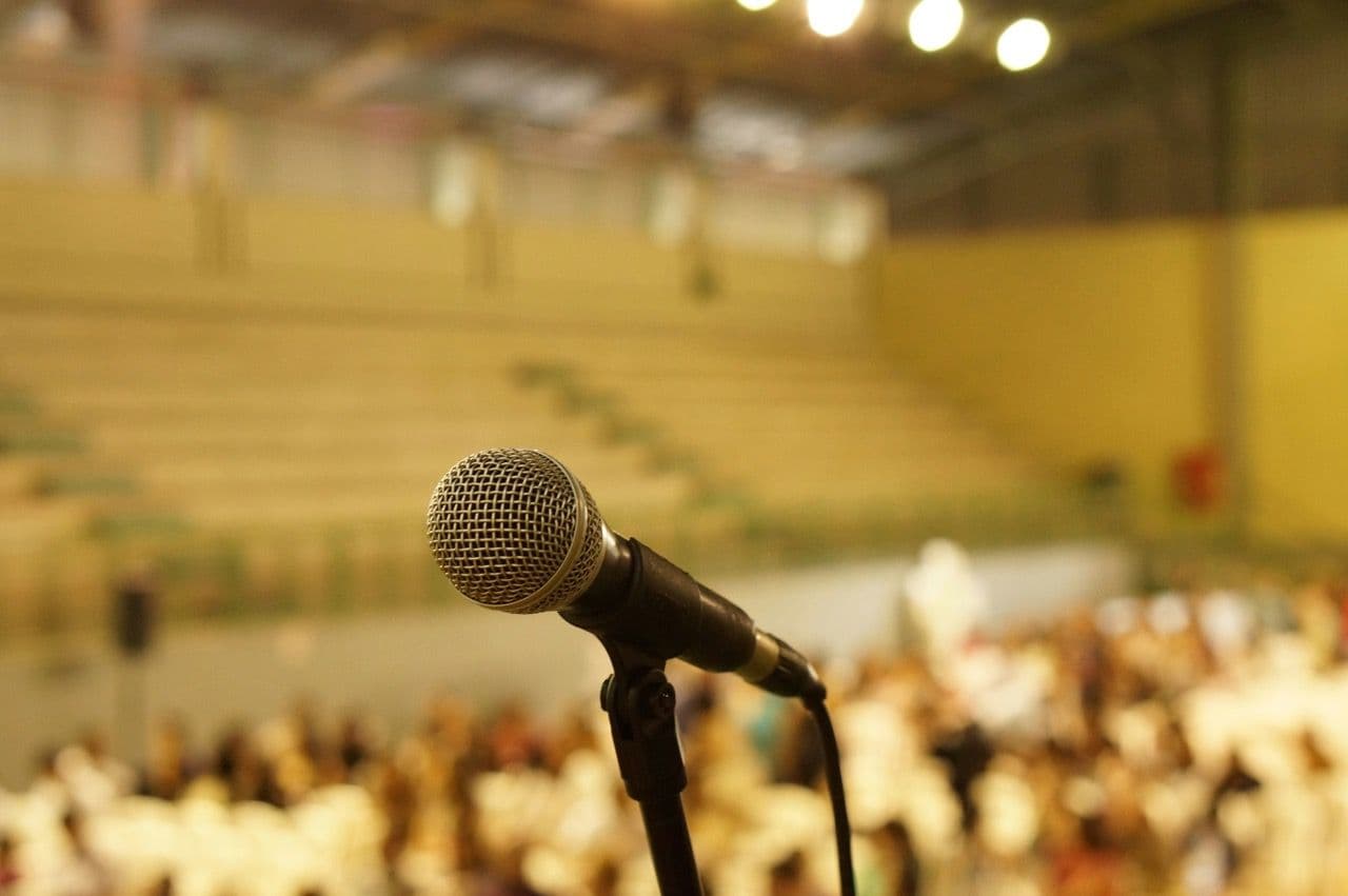 A microphone on a stand in focus, with a blurred large audience and tiered seating in an auditorium.