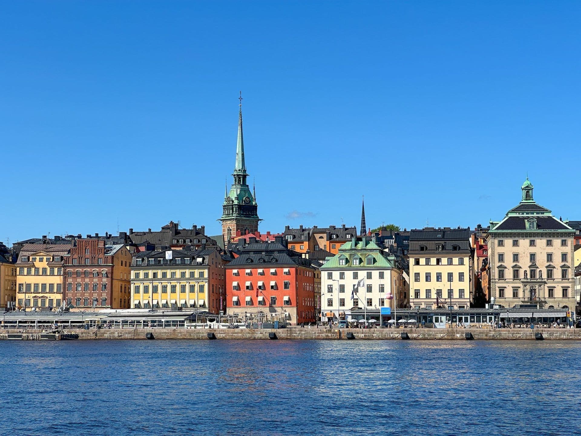 Historic colorful buildings and a prominent church spire line the Stockholm waterfront under a clear sky.