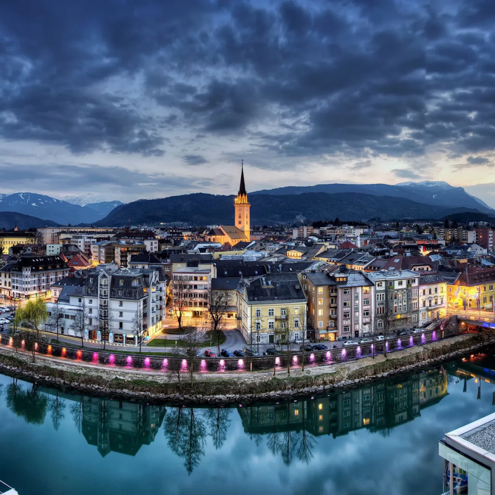 Villach, Austria, cityscape at dusk, showcasing a river reflecting illuminated buildings and mountains.